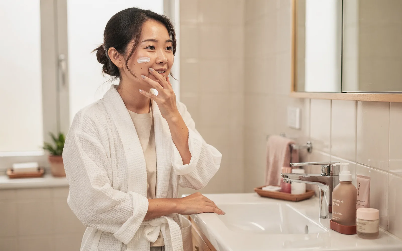 Woman in a white robe applying moisturizer at a bright, minimal bathroom sink.
