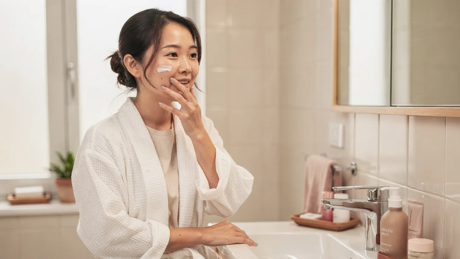 Woman in a white robe applying moisturizer at a bright, minimal bathroom sink.