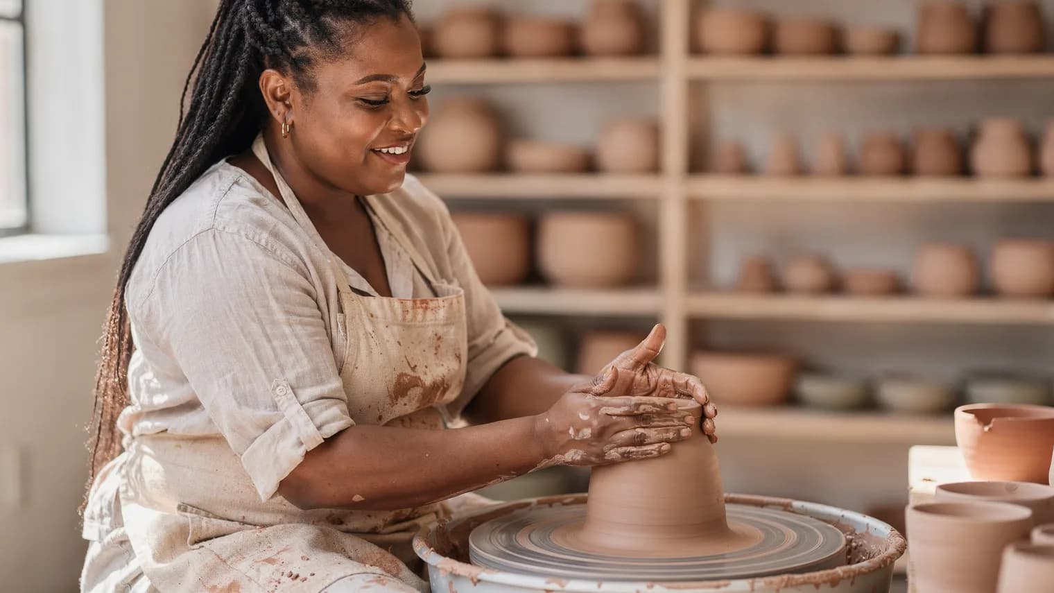 A woman in a clay-dusted apron shapes a vessel at a pottery wheel in a bright studio, absorbed in the work.