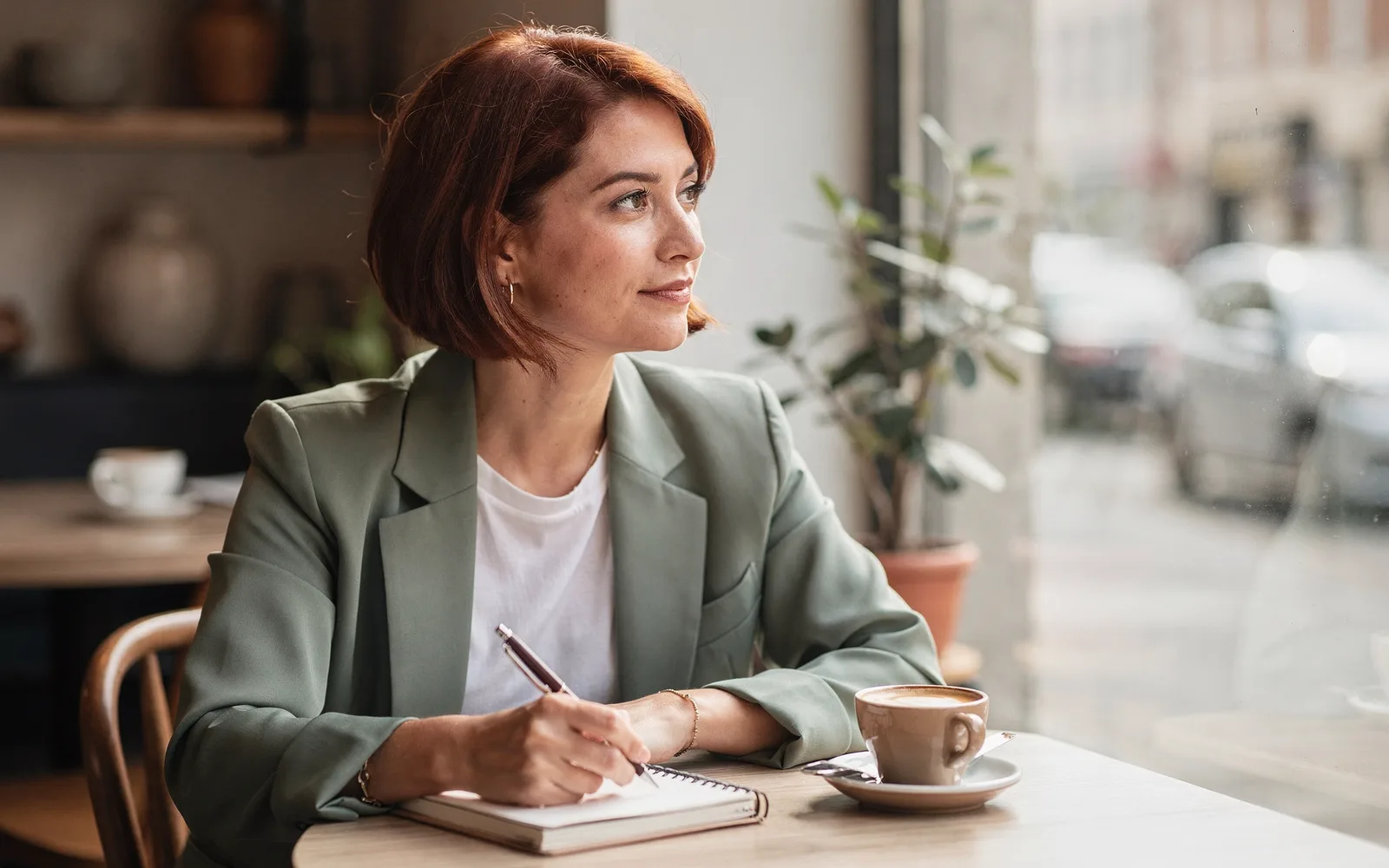 A woman at a café window table rests a pen on a closed notebook beside her coffee, looking out at the street.