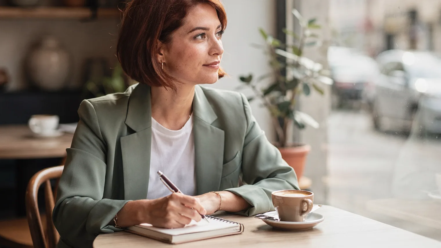 A woman at a café window table rests a pen on a closed notebook beside her coffee, looking out at the street.