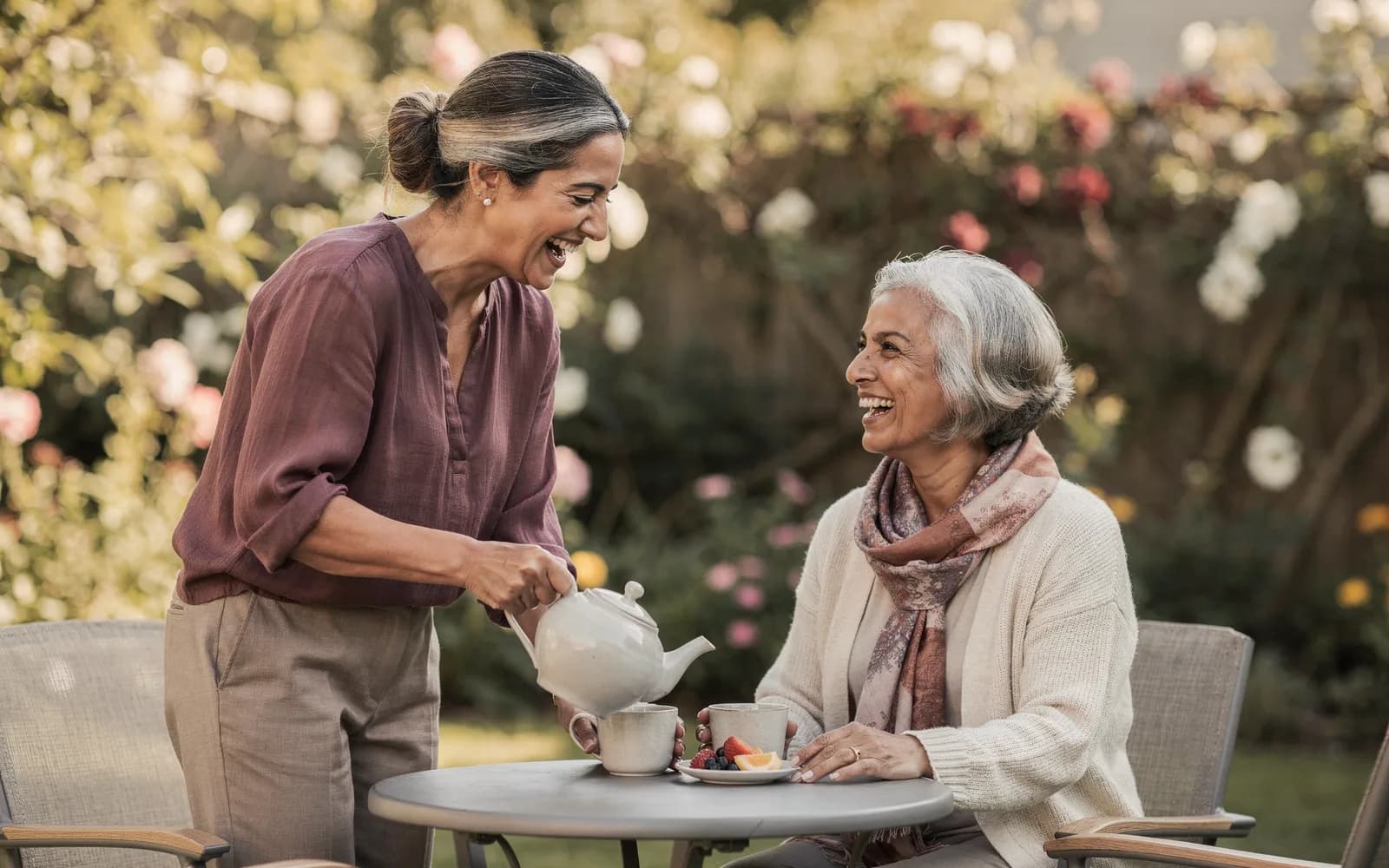 A woman pours tea for her elderly mother at a garden table, both laughing in dappled sunlight.
