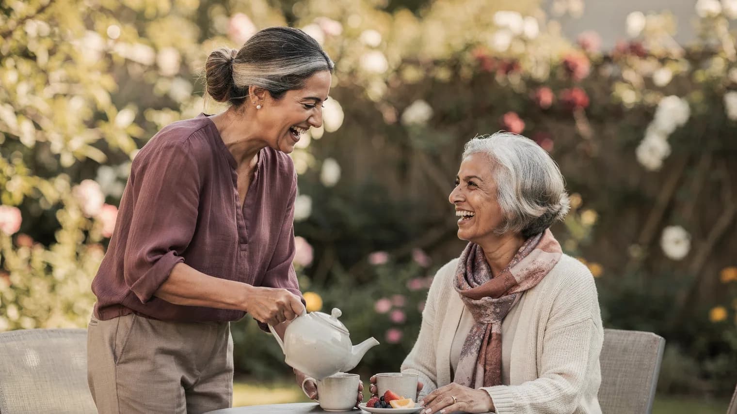 A woman pours tea for her elderly mother at a garden table, both laughing in dappled sunlight.