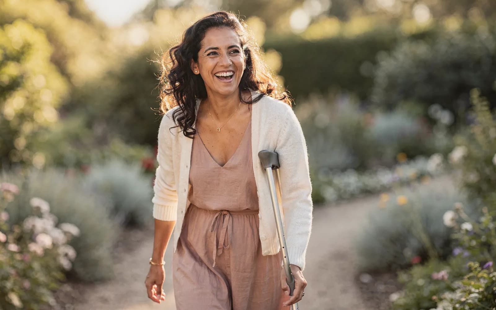Woman with loose curls laughing on a sunny, flower-lined garden path, walking with a forearm crutch.