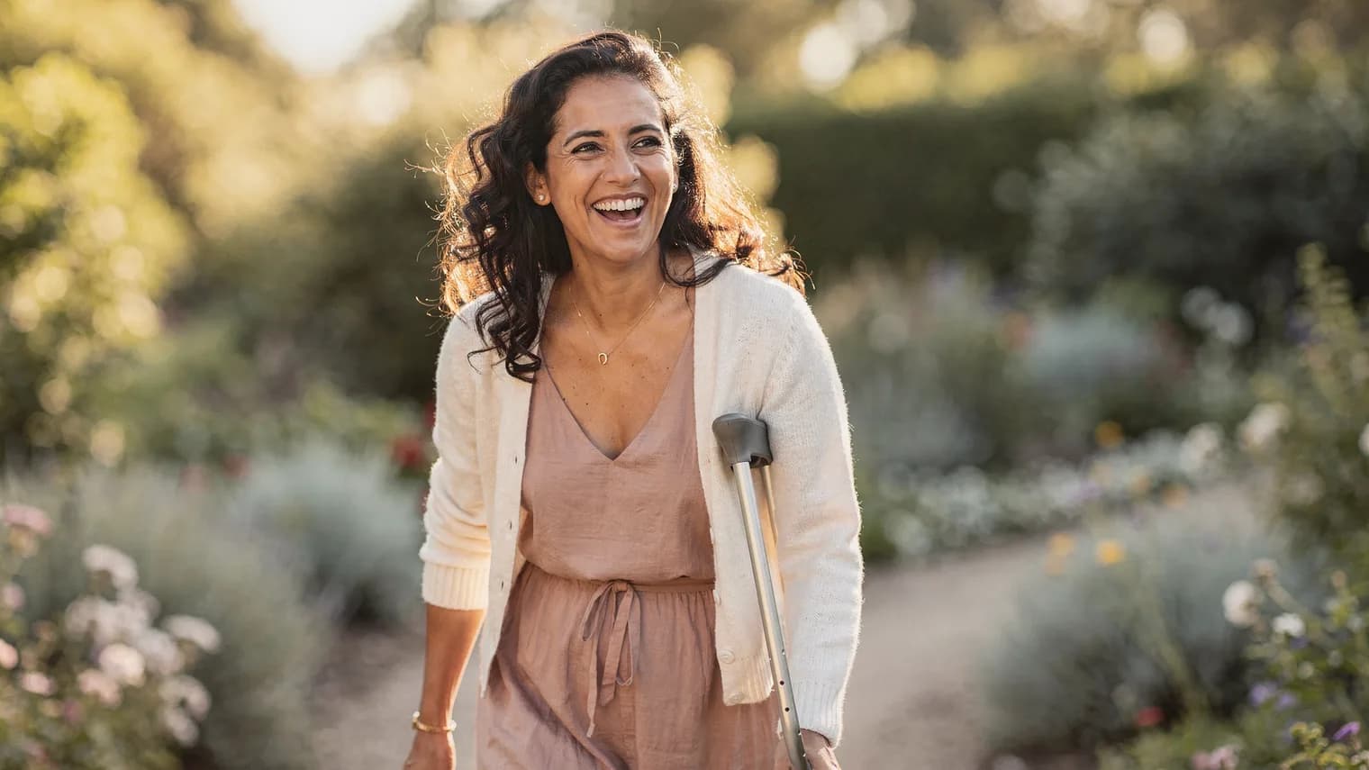 Woman with loose curls laughing on a sunny, flower-lined garden path, walking with a forearm crutch.