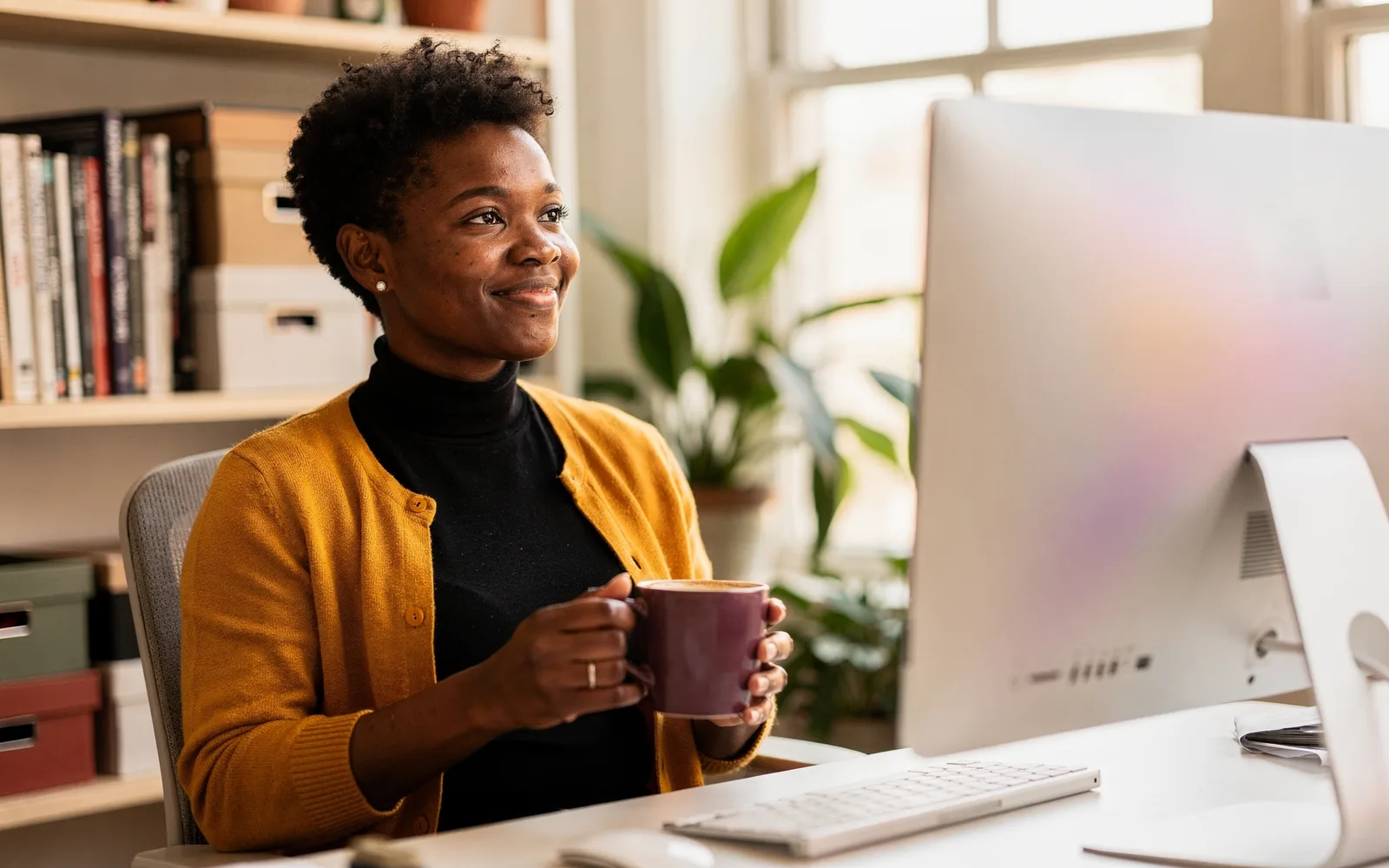 A woman with cropped hair sits at a studio editing desk with coffee, watching a softly glowing monitor with quiet pride.