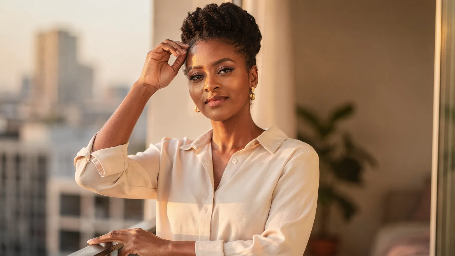 Woman with an elegant coily updo standing on a balcony at golden hour, hand resting near her hairline.