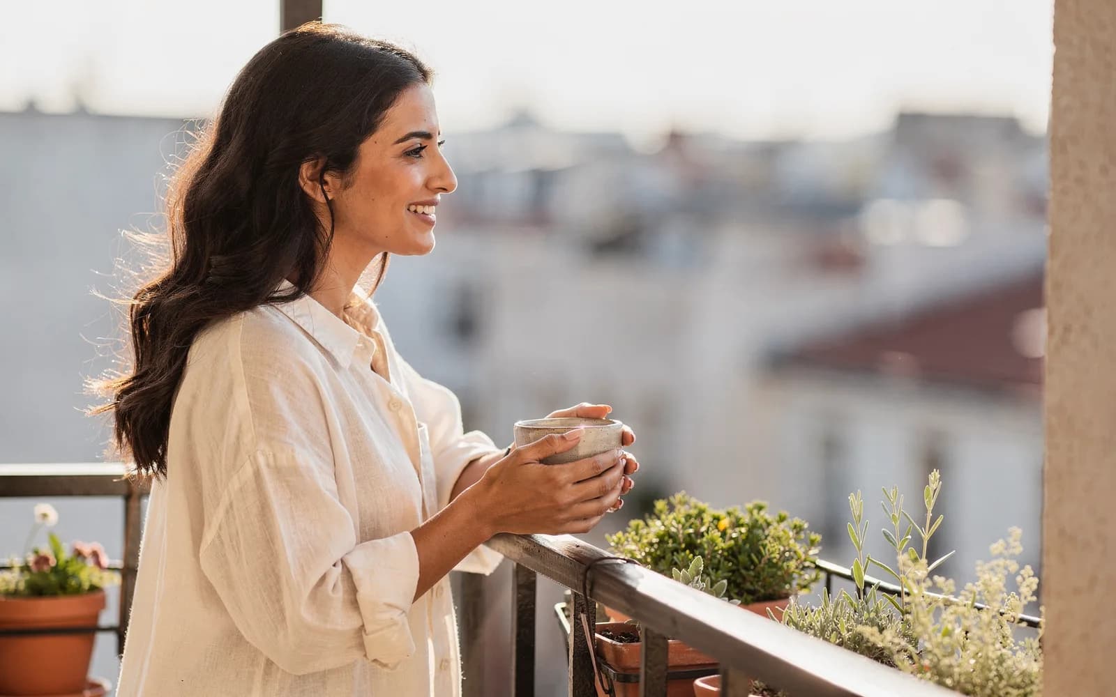 A woman in a linen shirt leans on a balcony rail with a cup of tea, looking calmly out over morning rooftops.
