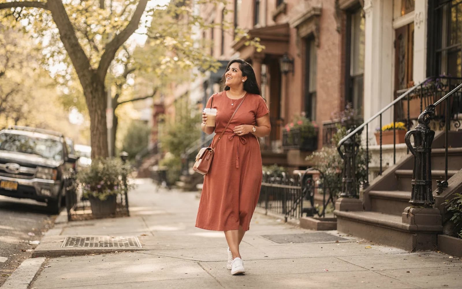A woman in a terracotta dress walks a tree-lined city street with an iced coffee, smiling slightly in golden light.