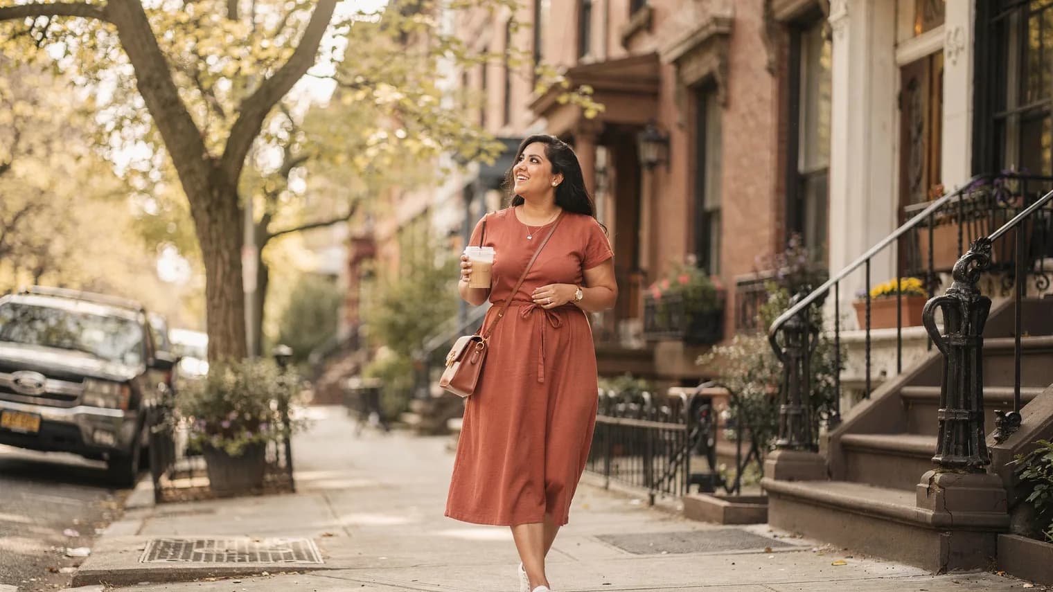 A woman in a terracotta dress walks a tree-lined city street with an iced coffee, smiling slightly in golden light.