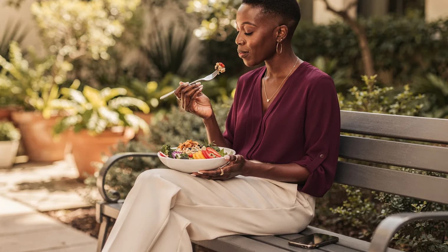 Woman enjoying a colorful grain bowl on a leafy courtyard bench at midday.