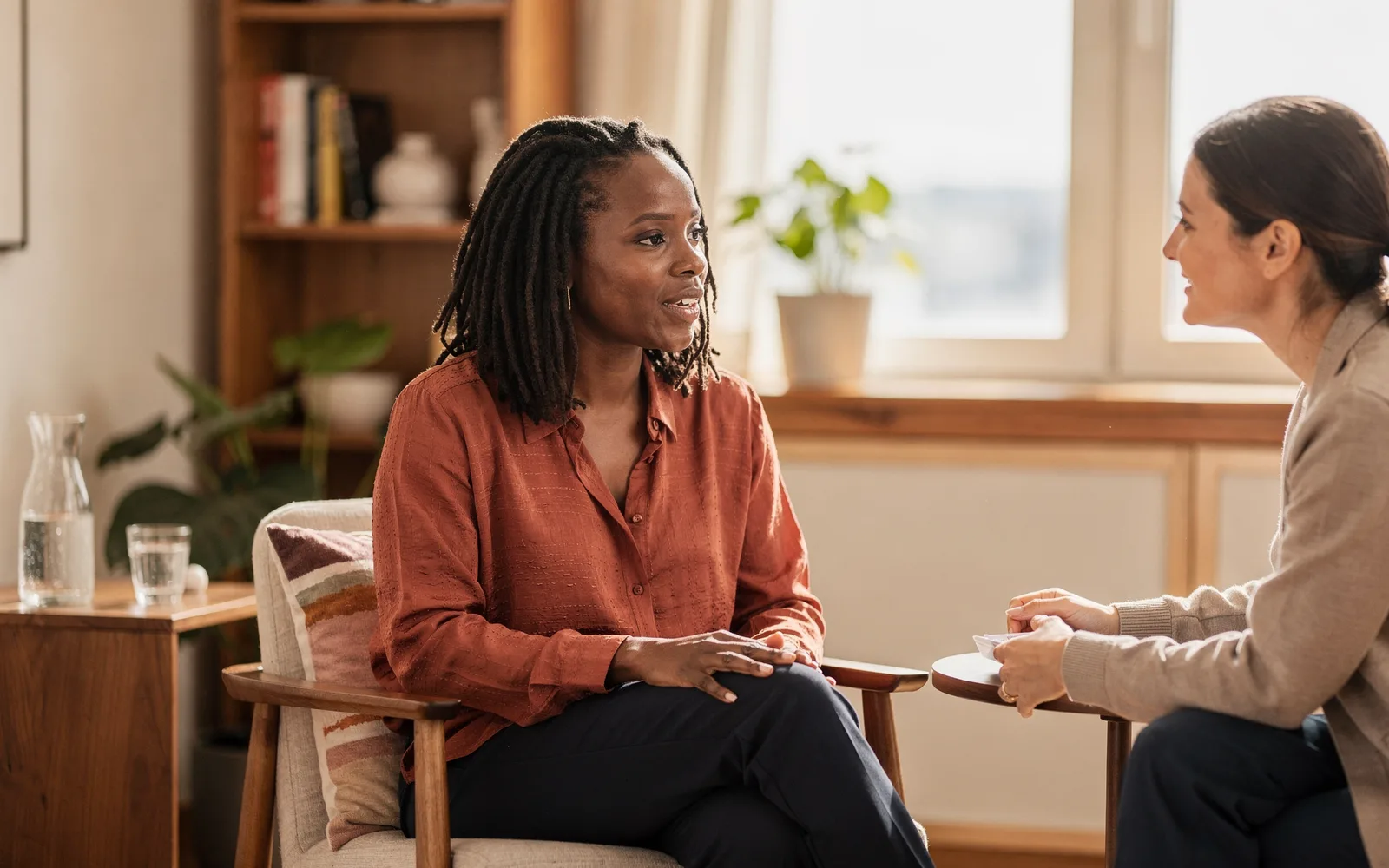 In a warm, plant-filled office, a woman speaks confidently while another woman across the table leans in and listens.