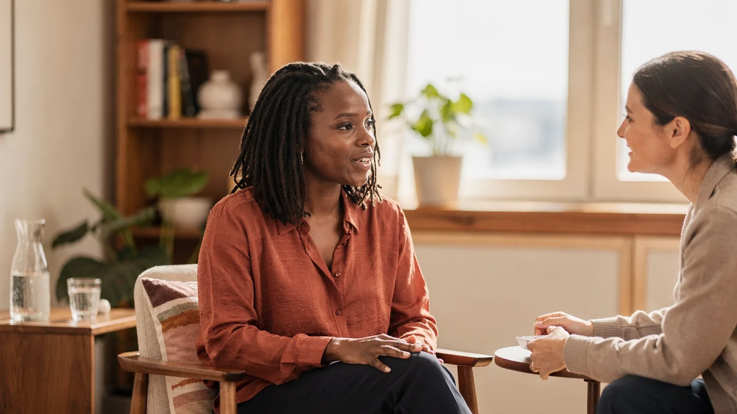In a warm, plant-filled office, a woman speaks confidently while another woman across the table leans in and listens.