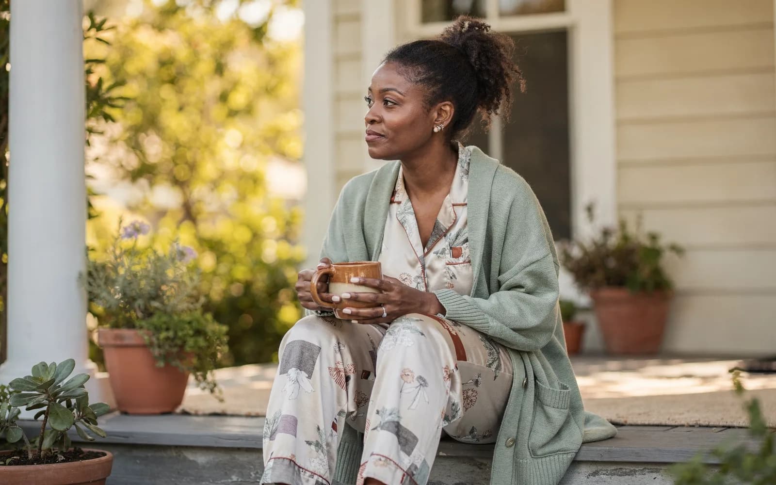 Woman in a sage cardigan sitting on a porch step with a mug in early-morning light.