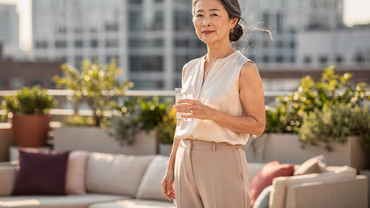 Woman in an ivory silk blouse holding iced water on a breezy planted office terrace.