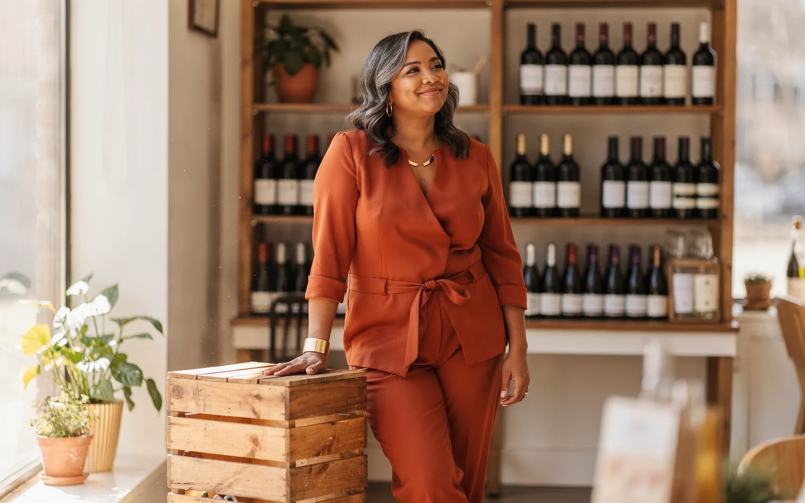 Woman in a rust-colored jumpsuit standing amused among wooden shelves in a small sunlit wine shop.