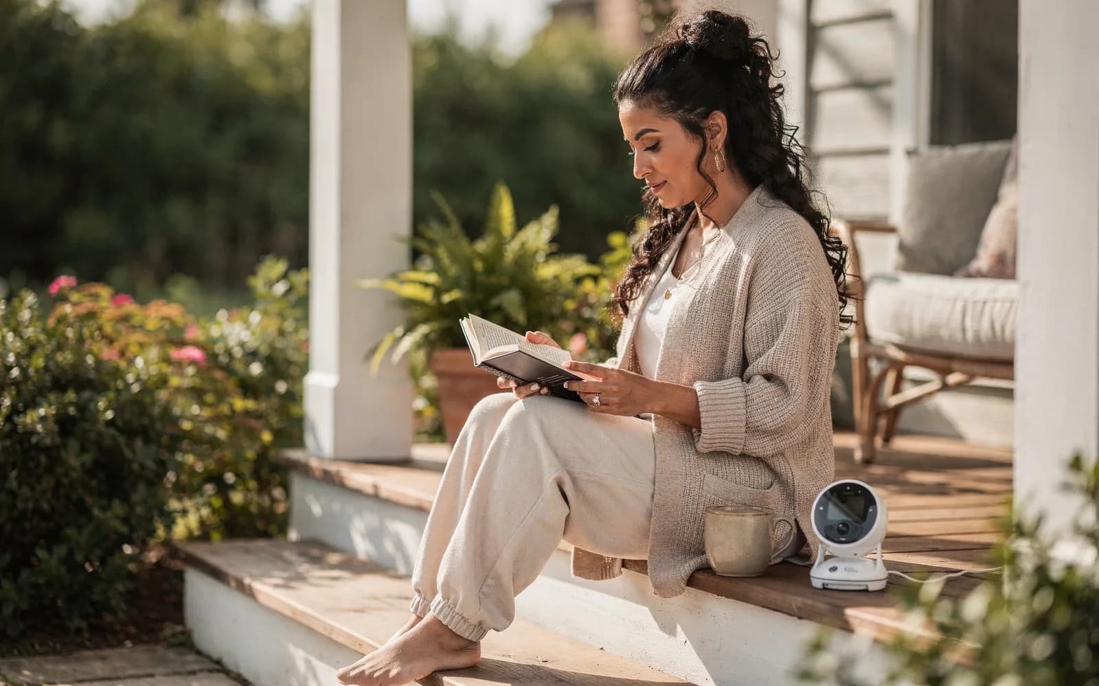 A woman reads a paperback on sunny porch steps with coffee beside her, relaxed in a morning moment of her own.