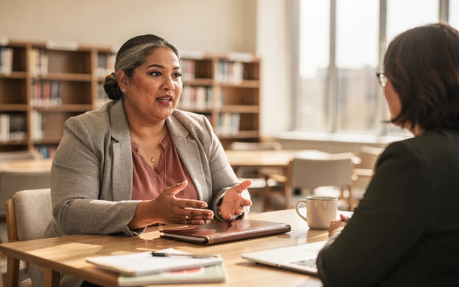 A woman leans forward confidently in conversation across a sunny library table, a closed portfolio between her and her companion.