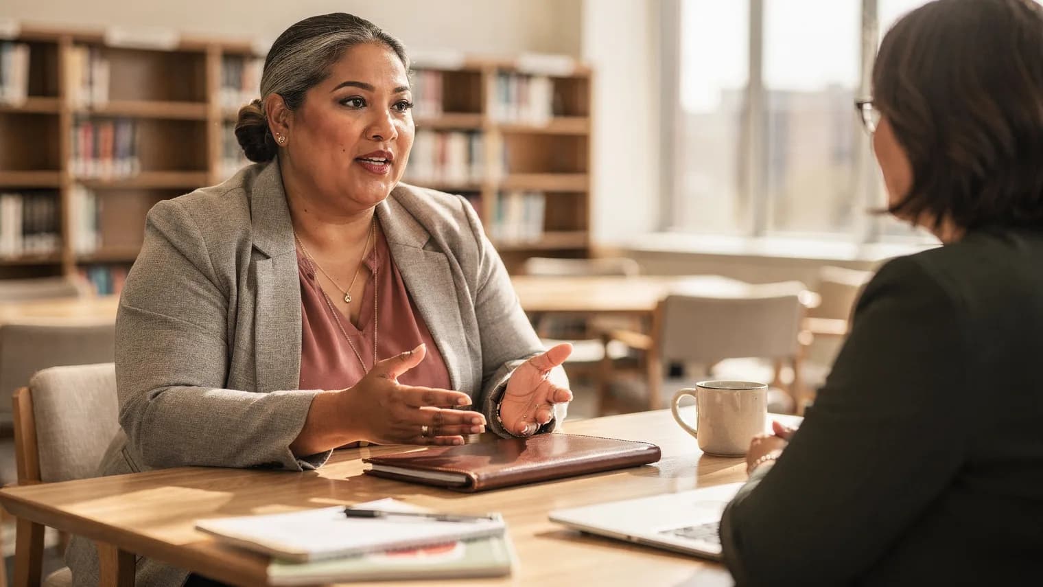 A woman leans forward confidently in conversation across a sunny library table, a closed portfolio between her and her companion.