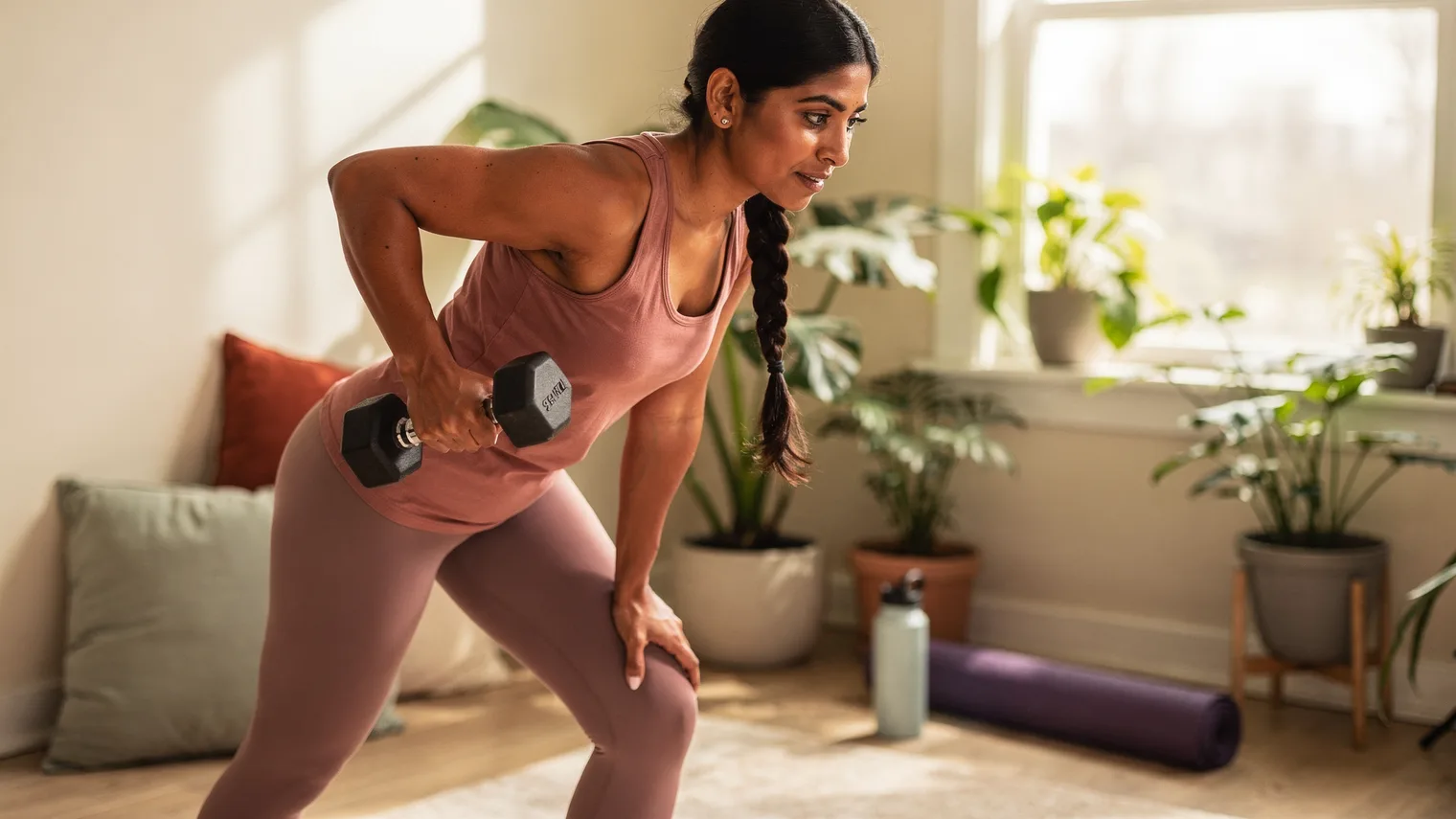 Woman doing a dumbbell row in a bright, plant-filled home workout corner.