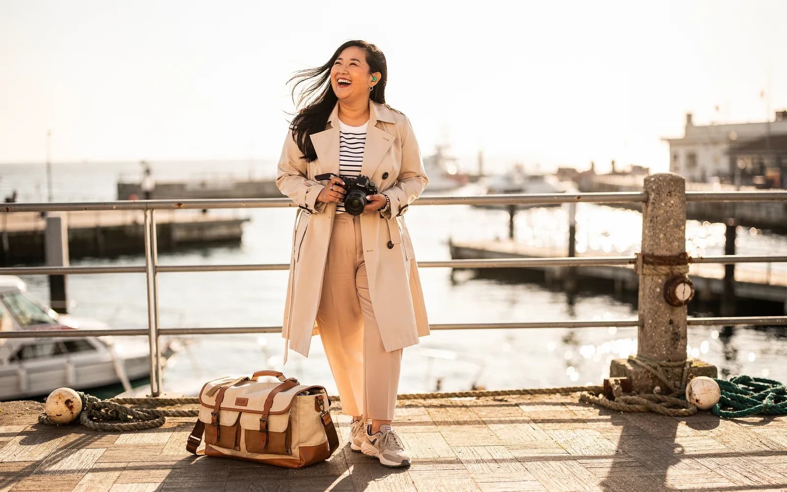 A traveler laughs into the wind at a harbor overlook, camera in hand and bag at her feet, water glittering behind her.