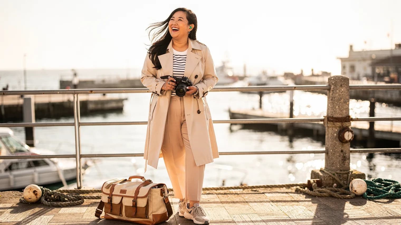 A traveler laughs into the wind at a harbor overlook, camera in hand and bag at her feet, water glittering behind her.