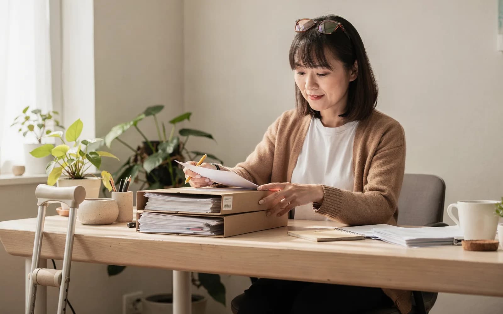 A woman at a sunlit home desk files documents into neat folders, plants around her and a crutch resting against the desk.