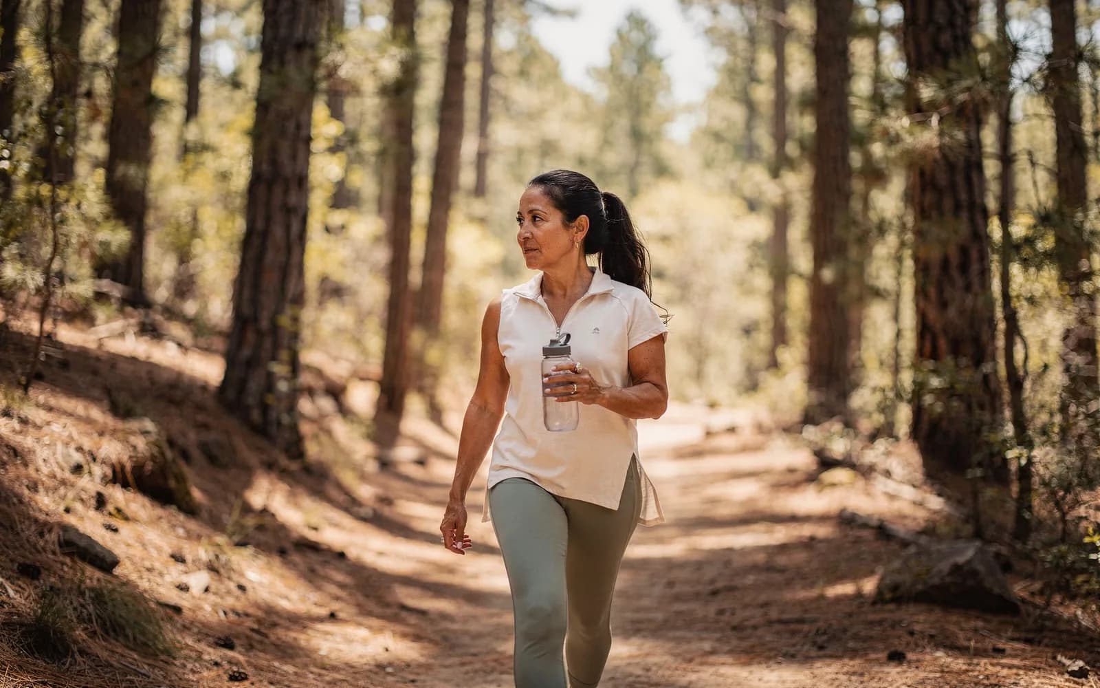 Woman in athletic wear walking a sun-dappled pine trail with a water bottle.