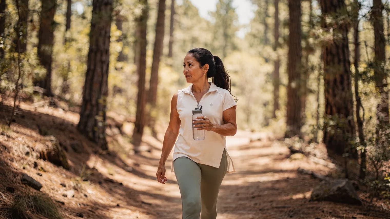 Woman in athletic wear walking a sun-dappled pine trail with a water bottle.