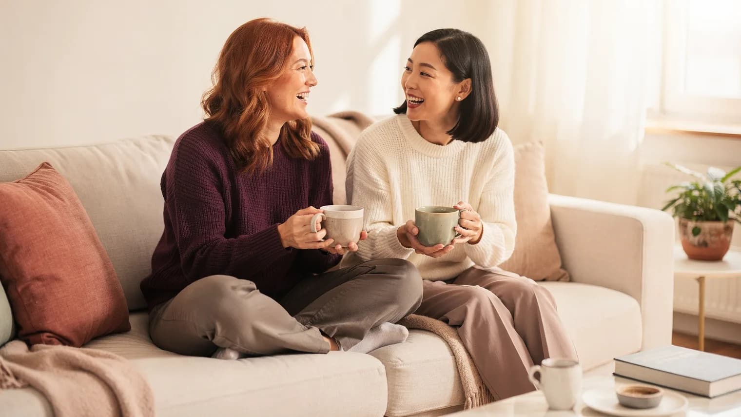 Two friends laughing softly over mugs of tea on a sofa in warm afternoon light.