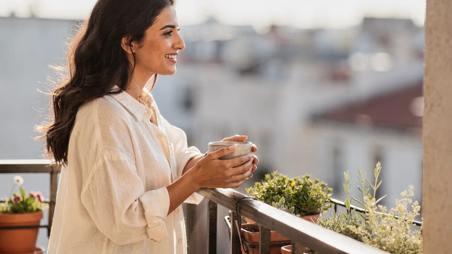A woman in a linen shirt leans on a balcony rail with a cup of tea, looking calmly out over morning rooftops.