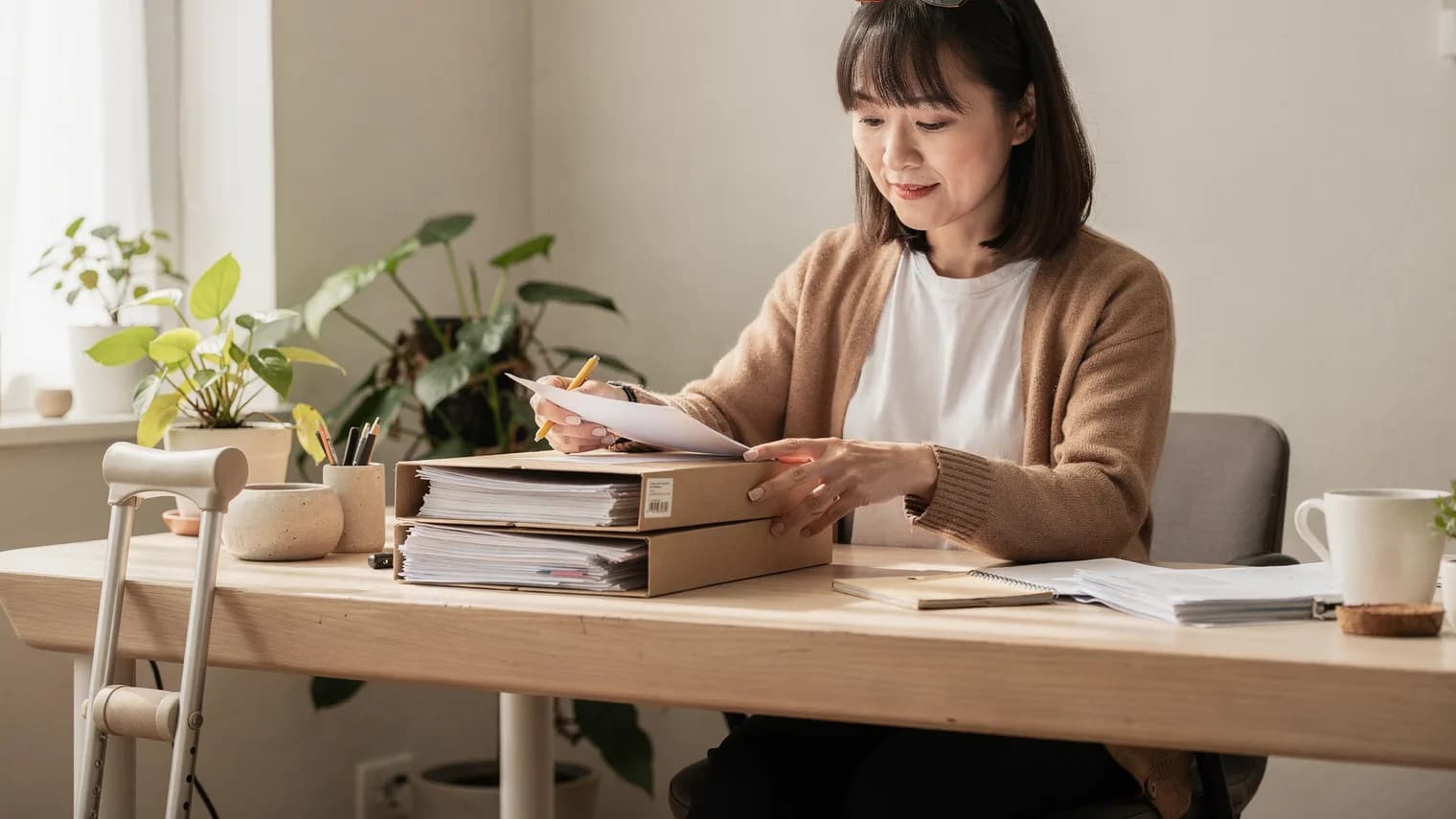 A woman at a sunlit home desk files documents into neat folders, plants around her and a crutch resting against the desk.